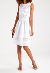Vestido blanco sin mangas con cintura ajustada y falda acampanada, que presenta un borde de encaje en el dobladillo y está hecho de algodón suave.