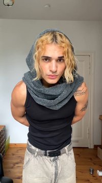 Black tank top, gray knitted scarf, short curly blonde hair, muscular arms, light gray jeans with studs, background with light wood flooring.