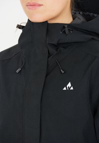 Svart vattentät jacka med hög krage, justerbara dragsko, och en vit logotyp. Materialet verkar slätt och hållbart.