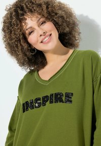 Sudadera verde de cuello en V con la palabra "INSPIRAR" en lentejuelas negras. Presenta un ajuste relajado y detalles desgastados en el escote y las mangas.