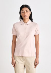 Polo shirt em rosa pálido com colarinho, mangas curtas e fecho de botão frontal. Apresenta um bordado discreto do logótipo no lado esquerdo do peito.