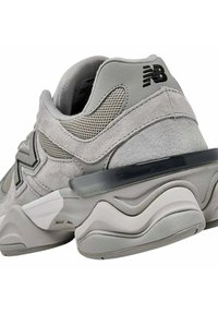 Scarpa sportiva grigia con tomaia in mesh e suede, suola curva, logo nero e accentuazione trasparente sul tallone. Presenta superfici testurizzate e un design a strati.