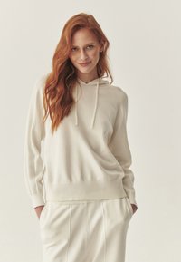 Kremowy sweter z kapturem, z prążkowanymi mankietami i dołem. Miękka tekstura, luźny krój, uzupełniony pasującymi spodniami. Prosty, minimalistyczny design.
