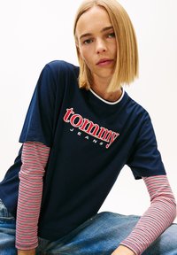 Granatowy t-shirt z czerwono-białym logo "tommy JEANS", noszony na czerwono-białych paskowanych długich rękawach. Luźny krój, materiał bawełniany.