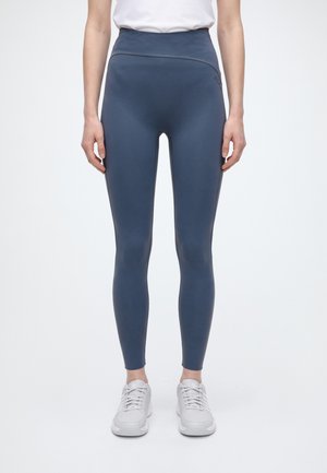 Modèle portant un legging bleu marine taille haute, un haut blanc et des baskets blanches sur un fond uni.