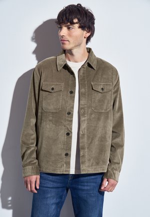 Chaqueta de camisa de pana verde oliva con dos bolsillos en el pecho, cierres de botón negros y un corte holgado, utilizada sobre una camiseta de color claro.