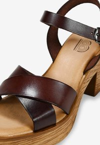 Sandal de cuero marrón con tiras cruzadas, tacón de bloque de madera y superficie texturizada. Correa ajustable con hebilla de metal.