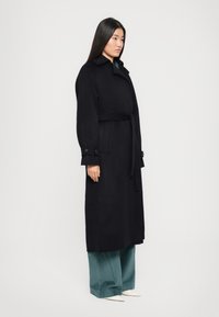 Garšs melns vilnas mētelis ar jostu viduklī, plašu apkakli un pogātām piedurknēm. Kombinēts ar platajām tirkīzzilajām korduroja biksēm un smailām baltām kurpēm.