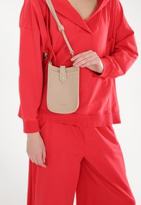 Beige leren crossbody tas met een gestructureerd oppervlak, rechthoekige vorm, drukknoopsluiting en gouden hardware, gedragen bij een rode outfit.