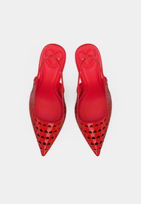 Chaussures slingback rouges à bouts pointus avec de petites découpes en forme de cœur et des motifs en forme de cœur sur les semelles intérieures, sur fond blanc.