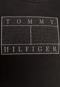 Tissu noir avec un contour rectangulaire blanc cousu contenant le texte "TOMMY" en haut et "HILFIGER" en bas à l'intérieur du rectangle.