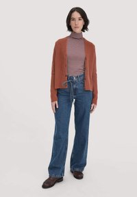 Ein rostfarbener Cardigan mit gerippter Textur, Knopfleiste, getragen über einem gestreiften Rollkragenpullover. Kombiniert mit weit geschnittenen Jeans und braunen Schuhen.