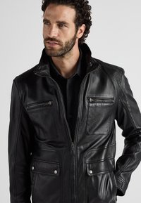 Schwarze Lederjacke mit durchgehendem Reißverschluss, zwei Brustreißverschlüssen, strukturierten Schulterakzenten und vorderen Klappentaschen. Glatte Oberfläche mit detaillierter Steppnaht.