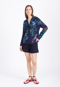 Une femme porte un cardigan boutonné bleu marine à fleurs, un short noir et des sandales rouges à lanières, debout devant un fond blanc uni.