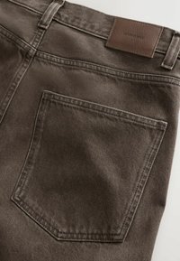 Közeli kép sötétbarna farmernadrág hátsó zsebének varrásáról, amelyen látható a cérnavarrás és a derékrészen egy bőr márkajelzés olvasható, melyen a "DENIM SUPPLY" felirat szerepel.