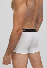 Boxer ajustados de algodón blanco con una cinturilla negra que presenta el logotipo "BOSS". Textura suave con un diseño ceñido, mostrando el perfil lateral y detalle del brazo.