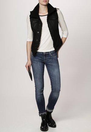 Femme portant une chemise blanche à manches longues, un gilet noir avec un col en fourrure, un jean skinny bleu, des bottines noires, tenant une pochette noire.