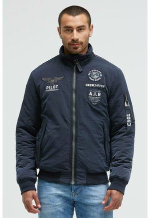 Navy bomber jacket made from synthetic material with a zip front, ribbed cuffs, and multiple patches including an eagle design.