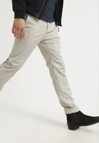 Persona che cammina indossando pantaloni beige, stivali neri alla caviglia, maglione blu navy e giacca nera su uno sfondo bianco.