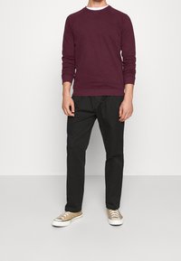 Mand iført en mørkerød langærmet sweatshirt, sorte bukser og beige sneakers, stående foran en ensfarvet hvid baggrund.