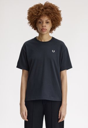 Mujer con cabello rizado y rojizo que lleva una camiseta negra lisa con un pequeño logo blanco en el pecho, de pie frente a un fondo claro y sencillo.