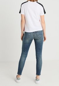 Camiseta blanca con acentos negros y rojos, jeans azules ajustados y zapatillas blancas con logo verde. Vista trasera, diseño casual, sin patrones visibles.