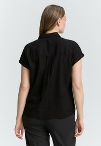 Chemise noire à manches courtes en tissu doux, avec un col boutonné et un détail de pli dans le dos. Le design est décontracté et légèrement ajusté.