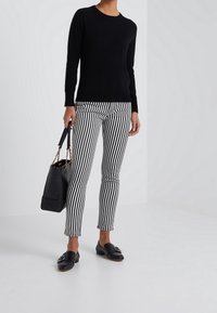 Schwarzer Pullover, gestreifte schwarz-weiße Hose, schwarze Handtasche mit goldenen Beschlägen und schwarze Slipper mit dekorativen Akzenten.