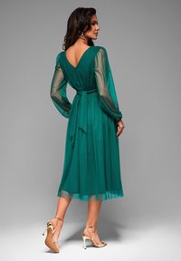 Teal midi-mekko, jossa on läpinäkyvät pitkät hihat, syvä V-mallinen selkä ja sidottava vyötärö. Yhdistetty kultaisiin remmillä varustettuihin korkokenkiin.