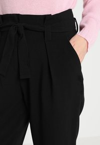 Pantaloni neri a vita alta con tasche laterali, cintura a fiocco e dettagli plissettati sul davanti, abbinati a un maglione rosa chiaro.