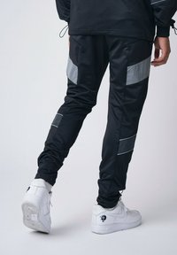 Pantalones deportivos negros con rayas y acentos grises reflectantes, puños ajustados y una textura suave. Combinados con zapatillas blancas.