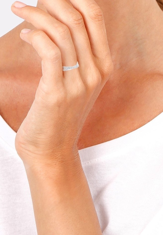 Main avec une fine bague en argent au doigt annulaire touchant doucement le menton, portant un haut blanc sur un fond neutre.