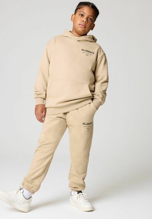 Beige Hoodie und Jogginghose Set mit Logodetail, lockerer Passform, aus Baumwollstoff und gerippten Bündchen. Kombiniert mit weißen Sneakers.