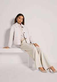 Vrouw in een lichtbeige jas, met brede broek en hakken, zittend in zijaanzicht op een witte bank tegen een effen achtergrond.