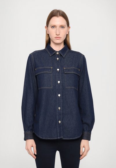 Chemise en denim bleu foncé avec un col classique, deux poches poitrine, et des boutons-pression en argent, à manches longues et avec des coutures contrastées.