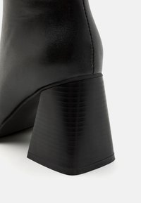 Black high-heeled boot with a tapered heel. The material appears smooth and shiny, with subtle stitching and a textured heel surface.