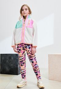 Veste en polaire de couleur menthe, rose et blanche, avec une fermeture éclair verticale ; associée à un legging imprimé léopard multicolore et des baskets beiges.