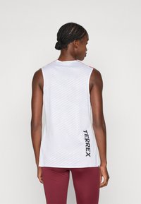 Débardeur de sport blanc sans manches avec un motif texturé, des accents rouges sur le col et "TERREX" imprimé en gras noir sur le côté.