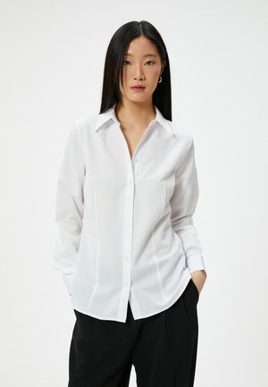 Camicia bianca con colletto, maniche lunghe e vestibilità slim. Il tessuto appare liscio e leggermente trasparente, rifinito con cuciture laterali.