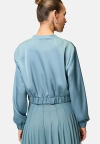 Helleblaue Satin-Bomberjacke mit elastischem Saum und Bündchen. Glatte Textur, leicht oversized Passform, schlichtes Design ohne Muster oder Beschläge.