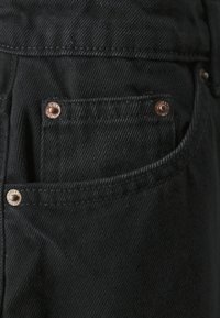 Jeans de mezclilla negros con un bolsillo delantero que cuenta con herrajes de bronce y costuras de contraste. La tela muestra una textura fina y un ligero brillo.