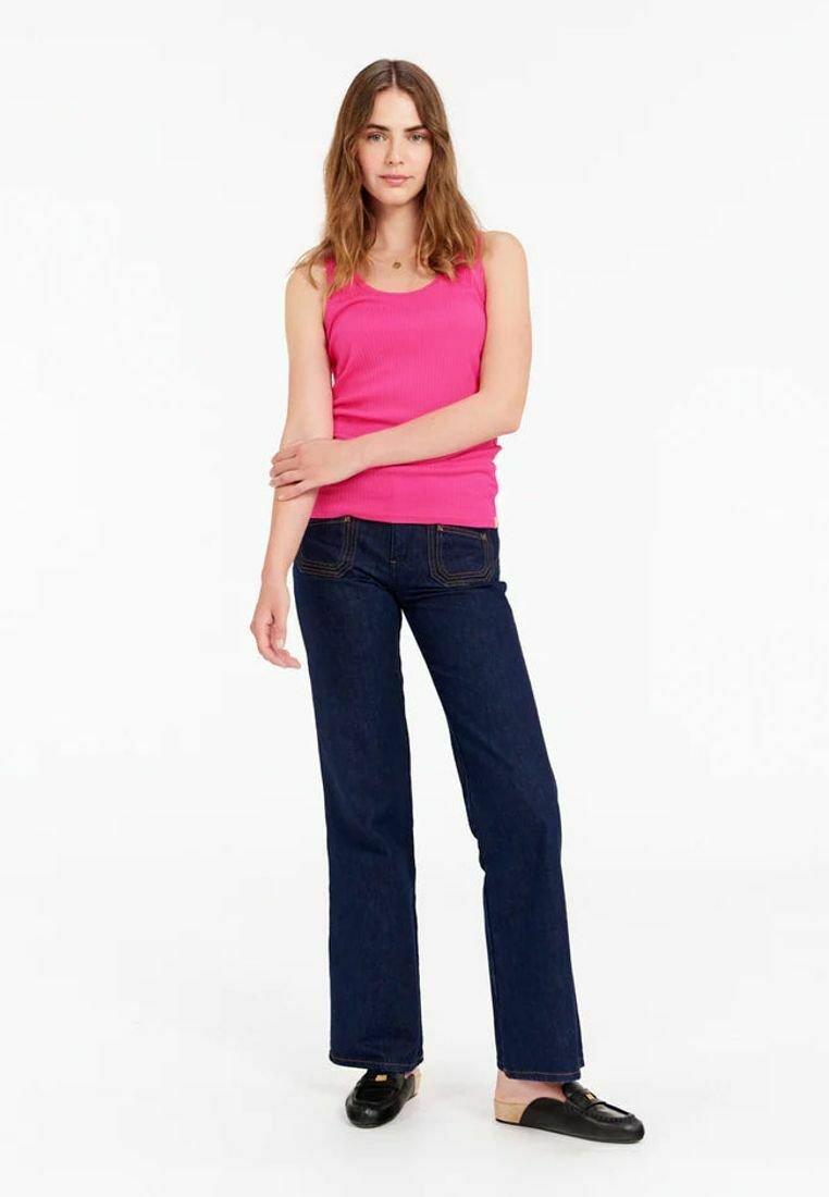 Top en rib à manches courtes rose, jeans évasés bleus foncés, chaussures noires à enfiler. Le mannequin se tient les bras croisés sur un fond blanc minimal.
