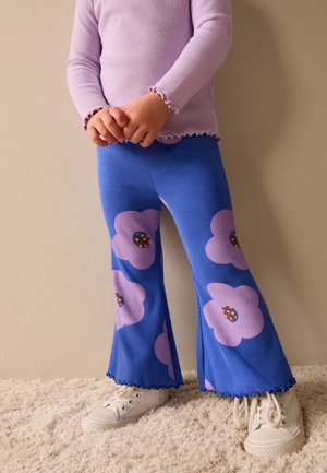 Copil cu pantaloni evazați albaștri cu flori mari mov, bluză lavandă cu mâneci lungi și pantofi sport albi, stând pe podea cu mochetă.