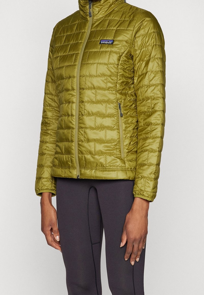 Personne portant une veste matelassée Patagonia vert olive à col montant et un pantalon noir, debout devant un fond clair et uni.