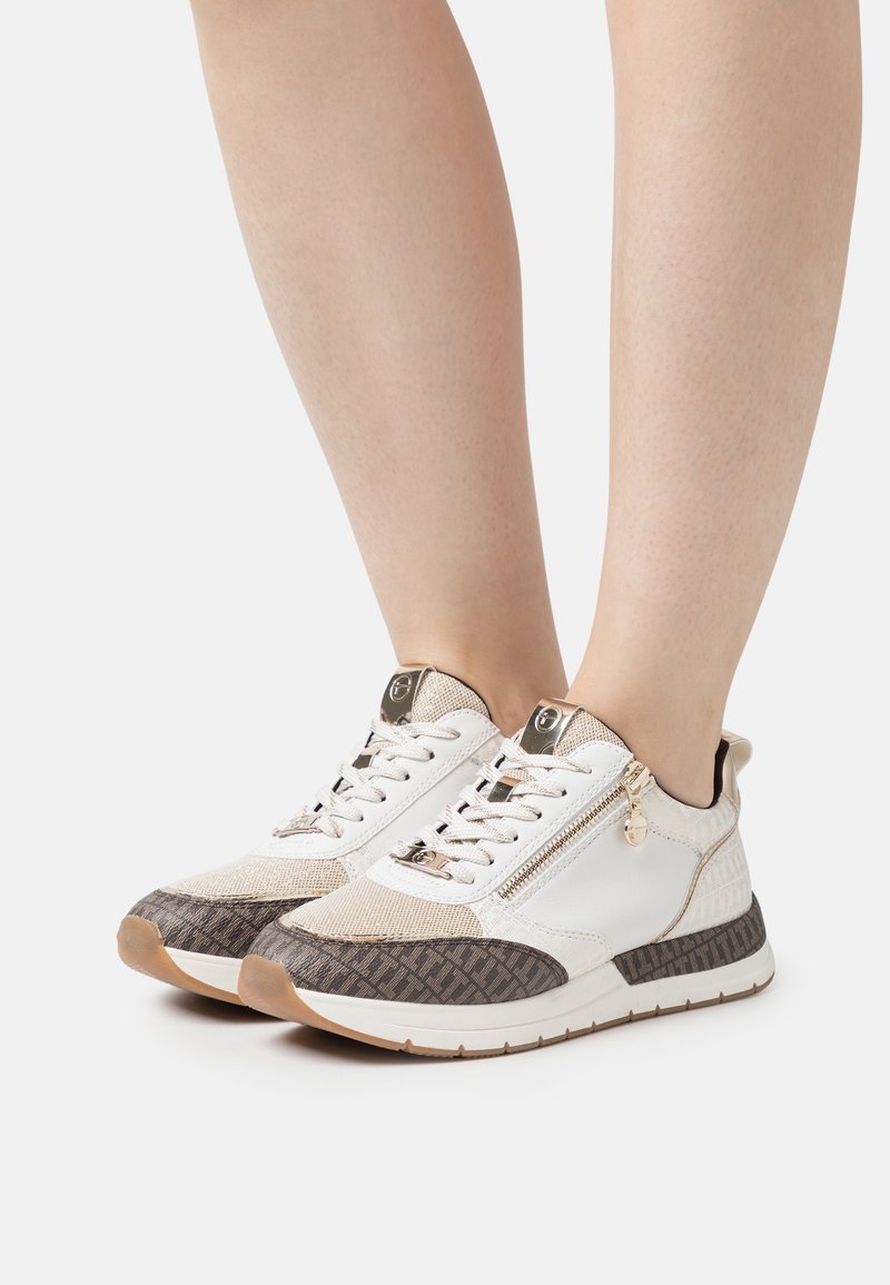 Jambes portant des baskets blanches et beiges avec des détails marron à motifs, des lacets et des fermetures éclair latérales sur un fond clair uni.