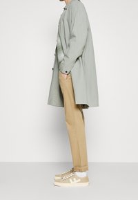 Cappotto lungo verde chiaro su una maglietta grigia a maglia, pantaloni beige e sneakers beige con accenti bianchi. Sfondo semplice.