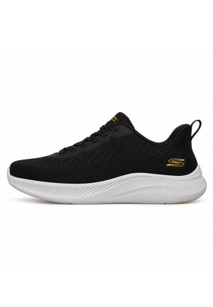 Zapatilla deportiva negra de punto con suela blanca texturizada, cordones negros y un logotipo amarillo en el talón y la lengüeta, mostrada de perfil lateral.