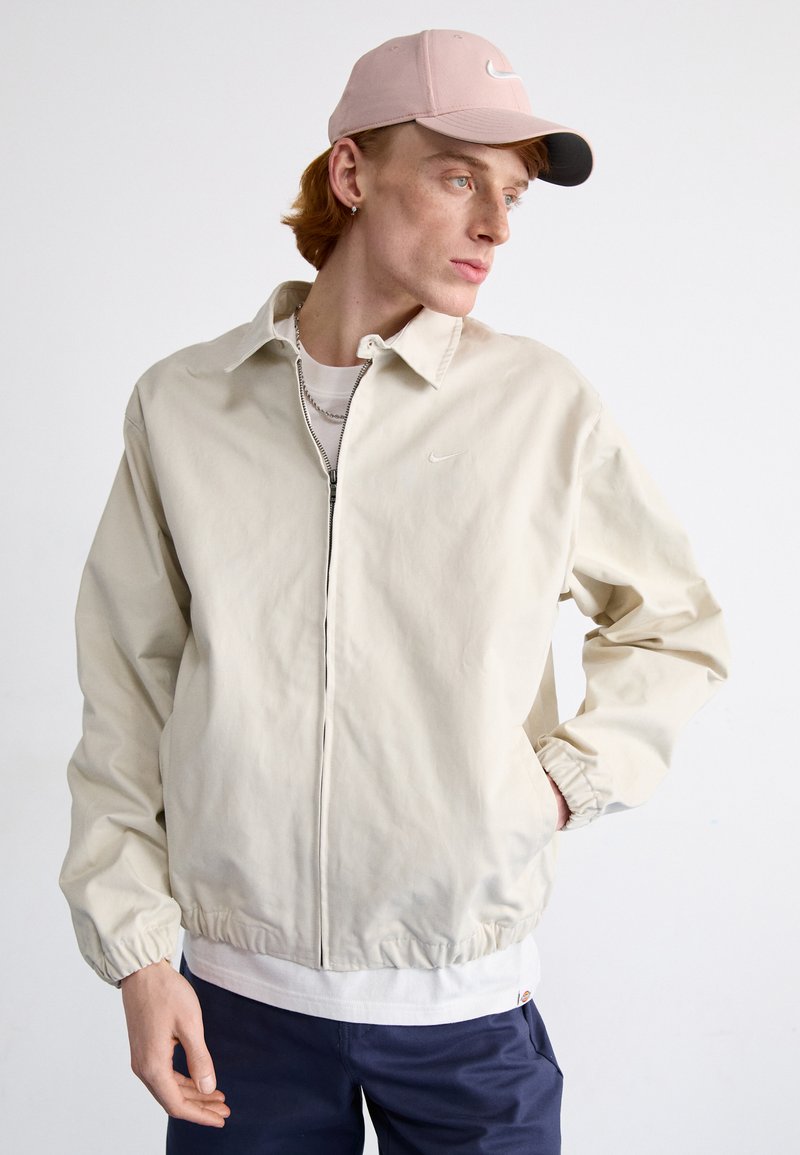 Lys beige zip-up jakke med krave, elastiske manchetter og talje; har et diskret Nike-logo på brystet og et lagdelt hvidt skjorte under.
