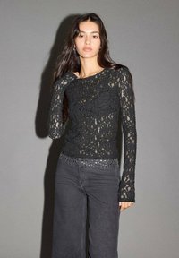 Crna čipkasta bluzica dugih rukava s teksturiranim uzorkom, u kombinaciji s tamnim trapericama visokog struka koje imaju srebrne zakovice duž pojasa.