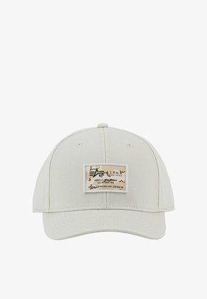 Șapcă bej deschis de baseball cu o etichetă dreptunghiulară pe care scrie „Alpha Industries, Knoxville, Tennessee, înființată în octombrie 1959” și detalii camuflaj.